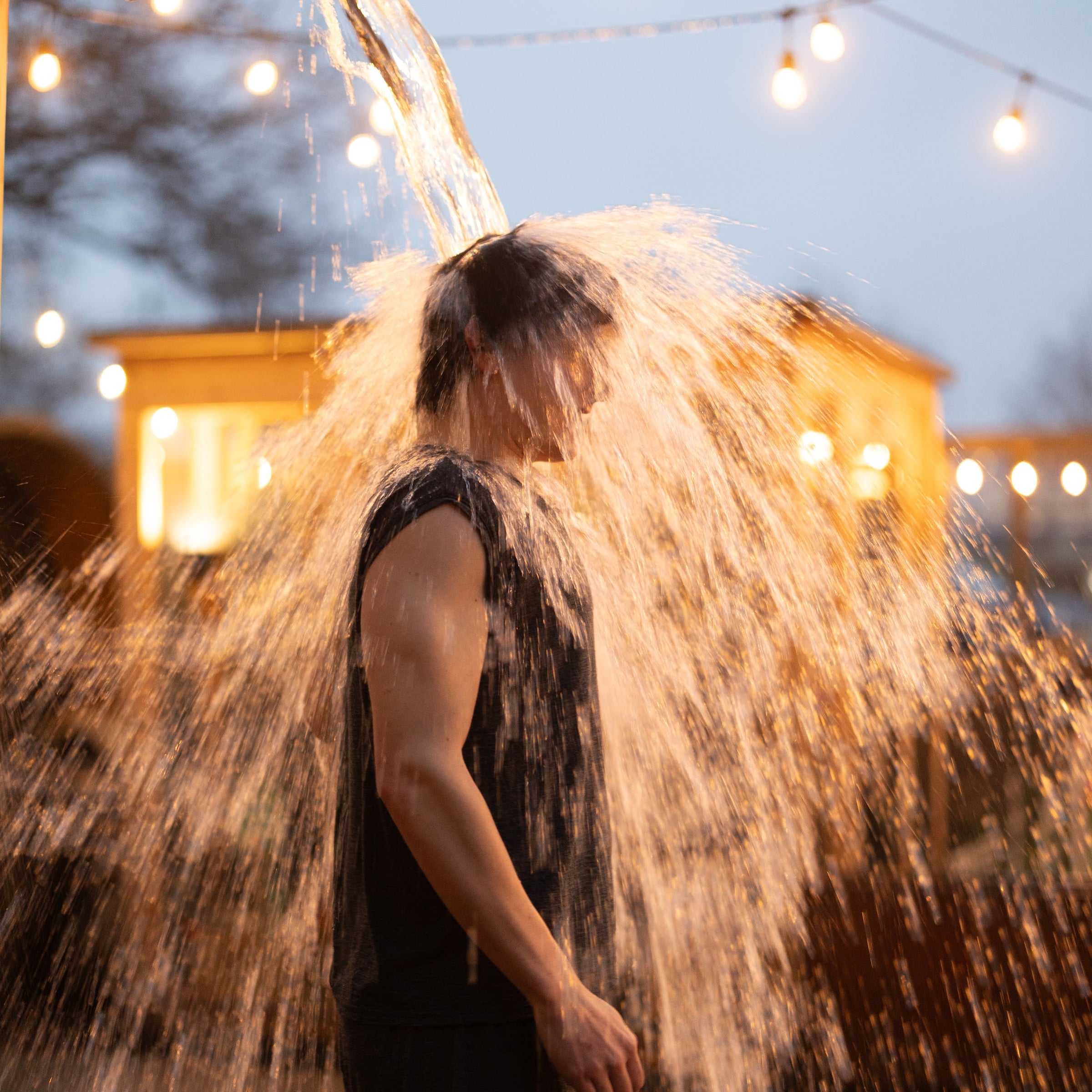 Person standing under a shower of water outdoors with string lights in the background